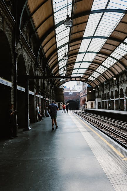 A railway station platform bordered by lush green trees with dense foliage and sunlight filtering through the leaves. The platform surface appears clean with a raised edge, and a metal gate at the end